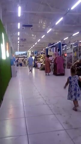 The video shows a brightly lit indoor marketplace or department store. The camera follows a young girl in a blue and white floral dress as she walks away from the camera down a wide aisle. She is likely walking towards her mother, who is pushing a stroller and is partially visible in the background to the right. Several other shoppers are scattered throughout the store, including a man in a light blue shirt walking towards the camera in the distance and a woman in a patterned dress browsing clothes on a rack. The store appears to have a high ceiling with exposed beams and numerous fluorescent lights. Merchandise is displayed on shelves and racks along the right side of the aisle, including clothing and various other items. On the left side of the aisle, a green, textured wall is decorated with illuminated signage that is difficult to decipher. The overall atmosphere is casual and appears to be a normal shopping day. There are no clear indicators of the time of day or weather, but the bright lighting suggests it could be daytime.