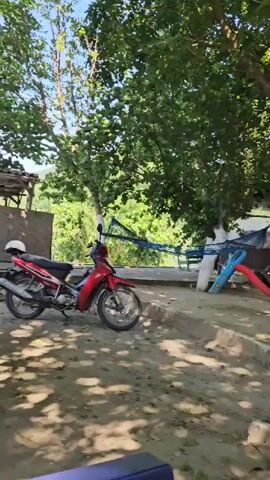 The camera pans right across a sunlit outdoor area with lush green trees. A red motorbike is visible on the left. Several wooden tables and benches are scattered across a dirt and concrete ground. In the background, there are low-rise buildings and what appears to be a blue and red playground slide. One tree trunk is painted white.