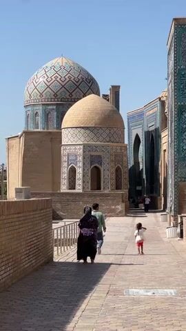 The video shows a sunny outdoor scene with several people walking along a paved pathway lined with ornate, ancient-looking buildings. The architecture features domes, intricate tilework in shades of blue, turquoise, and tan, and arched doorways. On the left, a woman in a dark, patterned dress walks away from the camera, followed by a man in a light green shirt. A young child in a white shirt and red shorts runs ahead of them. In the background, more people can be seen walking further down the pathway, and a bird flies in the clear blue sky. The overall impression is of a historical or religious site, possibly a mausoleum complex, in a warm climate.