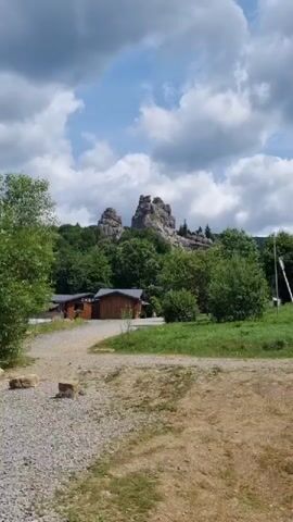 The video shows a natural outdoor scene. In the foreground, there is a gravel path leading towards a clearing with some dry vegetation. On the left side of the path, there are a few rocks. Further back, a small wooden building with a dark roof is visible. Behind the building, there is a dense area of green trees. In the background, prominent rock formations rise above the trees, under a sky with scattered white clouds against a blue backdrop. The overall impression is of a natural landscape with some man-made structures.