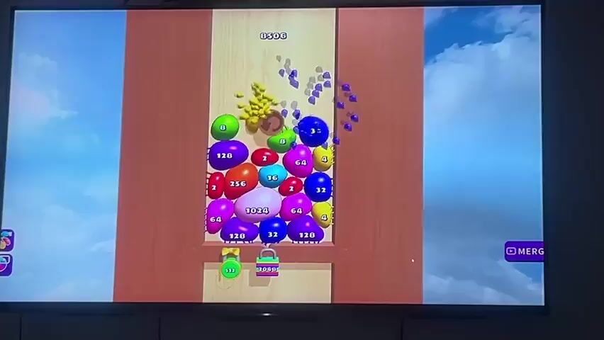 The video shows a close-up, medium shot of a television screen displaying a digital game. The game appears to be a puzzle or merging game with a vibrant, cartoonish aesthetic.

**Subjects:**
The primary subjects are the various colorful, numbered spheres and objects within the game's play area. These range in color from green, yellow, red, purple, blue, and pink. The numbers displayed on these spheres include 2, 4, 8, 16, 32, 64, 128, 256, and 1024, suggesting a progression or combination mechanic. There is also a brown, barrel-like object in the center of the play area, and two "locked" objects at the bottom, one green and one purple, with numbers 512 and 2000 respectively.

**Scene and Setting:**
The game's background is a sky with fluffy white clouds, suggesting an outdoor or ethereal setting. The play area itself is framed by wooden-textured borders, resembling an open door or window. The overall presentation is bright and cheerful.

**Activity or Event:**
The game involves dropping spheres from the top of the screen. The spheres are shown falling and accumulating in a pile. The goal of the game appears to be merging identical spheres to create higher numbered spheres. The video captures a sequence of spheres falling and the subsequent merging and rearrangement of the existing spheres within the play area. The score at the top of the screen shows a progression from 8534 to 8562.

**Notable Details:**
The game is being played on a modern flat-screen television. The lighting in the room where the television is located is not visible, but the screen itself is well-lit. There are no apparent environmental cues like time of day or weather, as these are depicted within the game's virtual environment. The emotions conveyed are those of a casual gaming experience, with bright colors and clear objectives.

**Visible Text:**
Within the game interface, the following text is visible:
* Numbers on the spheres (2, 4, 8, 16, 32, 64, 128, 256, 1024, 512, 2000)
* Score at the top of the screen (e.g., 8534, 8560, 8562)
* A button labeled "MERGE" on the right side of the screen.
* A brand name "SONY" below the TV screen.