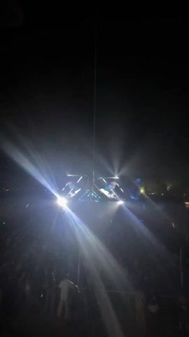 Here's a description of the video:

The video shows a concert stage at night, heavily illuminated by bright blue spotlights. The lights create intense flares, almost obscuring the performers on the screens above the stage. A large crowd is visible in the foreground, silhouetted against the bright stage lights. A thin wire or cable stretches vertically from the stage area into the dark sky above.