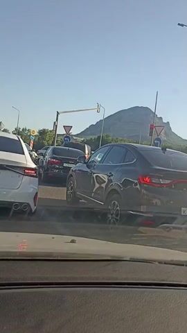 The video shows a traffic jam on a sunny day, likely in Mineralnye Vody, Russia. The camera is inside a car, capturing the view from the driver's perspective. In the foreground, the hood of the car and its dashboard are visible. Ahead, several cars are stopped on a multi-lane road. The closest car on the left is white, and a black car is to its right, in the next lane. Further ahead, more cars are visible, including a black sedan with a license plate starting with "H 291 XO". The traffic appears to be backed up at an intersection, indicated by traffic lights and road signs. One sign shows a red yield triangle, and another indicates a left turn.

In the background, a large, rugged mountain dominates the horizon under a clear blue sky. The sunlight suggests it is daytime, likely late afternoon or early evening given the angle of the light on the mountain. The scene is typical of urban or suburban traffic congestion. There are no visible people outside the vehicles. The mood is calm, as indicated by the stillness of the traffic and the clear weather. The visible text includes the license plate "H 291 XO" and partial text on a dark barrier in the background which includes the word "TRANSPORTS".