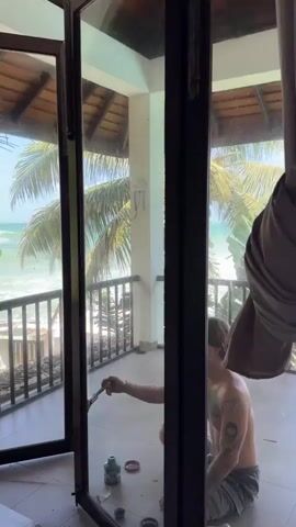 A young, shirtless man with tattoos on his arms and chest kneels on a tiled balcony, cleaning a large glass sliding door with a brush. He is wearing grey shorts. There are two green bottles and a small silver container on the floor near him. The balcony overlooks a tropical beach with palm trees, a railing, and the ocean under a bright, partly cloudy sky. The sun is shining.