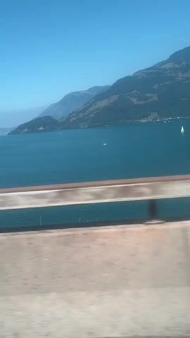 The video shows a scenic view from a moving vehicle, possibly a train or car, looking out over a large body of water. The water is a deep blue and appears calm, with a few small white sailboats dotting the surface. In the background, rolling green mountains rise up, their slopes covered in lush vegetation. The sky is a clear, bright blue, with a few wispy white clouds. The foreground is dominated by a railing or barrier, partially blurred due to the motion of the vehicle, and a concrete structure that appears to be part of a bridge or overpass. The overall impression is one of a peaceful and picturesque landscape on a sunny day.