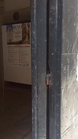 The video shows a close-up view of a dark, heavy-looking door. The door appears to be made of metal and has a weathered, rough texture with visible rivets and some signs of rust. The door is partially ajar, revealing a gap in the middle. A small padlock is visible securing the door. In the background, through the gap, a white poster or sign with text and images is visible, attached to a wall. The poster appears to be promotional, possibly for a tourist attraction or event. The lighting suggests an outdoor or semi-outdoor environment, possibly a passageway or entrance. The camera focuses on the door's handle and then pans to the lock. The sound includes the creaking of the door and a voice speaking in Italian, presumably describing the door and the action of knocking.