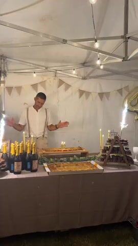 A man in suspenders stands behind a table laden with food and drinks, facing the camera with his hands on his chest. The table displays bottles of champagne, pastries, cupcakes arranged on a tiered stand, and candles. A large rectangular cake with lit candles sits on the table, and a sparkling decoration is visible on the left. The scene is set outdoors under a white tent adorned with string lights and triangular flags.