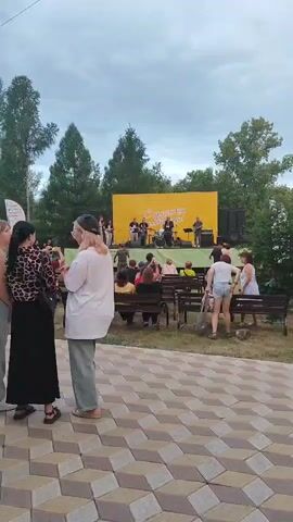 A band plays on a stage in front of a yellow backdrop. A crowd of people watch from benches and standing positions. The scene takes place outdoors, likely in a park, with trees visible in the background and a paved walkway in the foreground. The sky is cloudy. The video is recorded from a distance, making it difficult to discern specific details about the individuals or the band members.