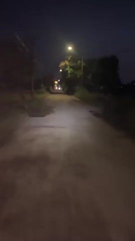 The video shows a person riding a bicycle at night down a poorly maintained asphalt road. The scene is dimly lit by spaced-out streetlights that cast a warm, yellow glow. On either side of the road are trees and foliage. Power lines are visible against the dark sky. There are potholes and uneven patches on the road surface. In the distance, car headlights are visible, approaching from the opposite direction. The bicycle's shadow appears briefly on the road.