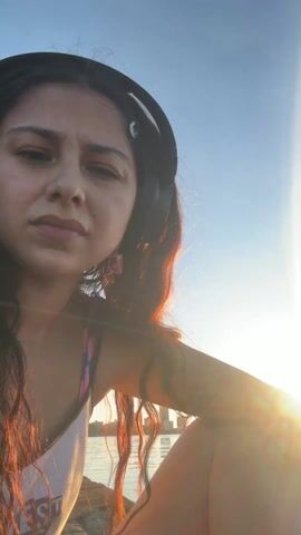A woman wearing headphones is outside and appears to be near a body of water. She says in Spanish that a spider bit her and that she thinks she will die. She shows her foot and looks concerned. The sun is shining brightly.