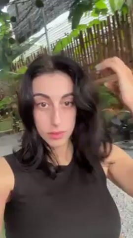 A woman with dark hair, wearing a black tank top, is seen in a selfie-style video. She is adjusting her hair with her hands. The background appears to be an outdoor setting, possibly a patio or garden, with greenery and wooden structures visible. The ground is covered in gravel. There are folding chairs and a fan nearby.