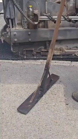 The video shows a close-up of an asphalt paving operation. In the foreground, a large, flat metal tool with a wooden handle, likely a lute or rake used for smoothing asphalt, rests on the ground. Behind it, a portion of a paving machine is visible, dispensing a layer of hot asphalt. Two individuals, dressed in work clothing, are present. One person, wearing work boots and tan pants, is partially visible on the left. Another person, wearing dark work pants with reflective stripes and an orange shirt, is more prominently seen on the right, with their back to the camera. The scene appears to be outdoors on a paved surface, likely a road construction site. The asphalt is fresh and dark, indicating it has just been laid. The lighting suggests it is daytime. No specific time of day or weather conditions are discernible. There is no visible text in the video. The context indicates the location is Hévíz, Hungary, consistent with roadwork activities. The overall atmosphere is one of industrial labor and construction.