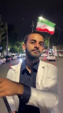 A young man with dark hair and a beard stands in the middle of a street at night, holding a flagpole. He is wearing a white lab coat over a dark blue shirt and black pants. The lab coat is open, revealing the shirt. He is holding the flagpole with both hands, and it is angled upwards to his right. A flag, appearing to be the flag of Iran, is attached to the top of the flagpole and is waving in the breeze. To his right, another flag, mostly yellow with some green, is also attached to the flagpole and is also waving. The street is paved and has lane markings. On either side of the street are buildings and trees. There are several cars driving on the street, some with their headlights on, illuminating the scene. A street lamp is visible in the background, casting a bright light. The overall impression is that the man is participating in some kind of public event or demonstration at night.