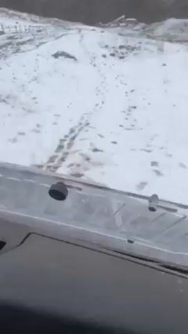 The video shows a vehicle driving on a snow-covered path with mountains in the background. The mountains are partially covered in snow and clouds. The path is lined with a wooden fence on one side. There are footprints in the snow on the path. The vehicle appears to be a utility terrain vehicle (UTV) or similar off-road vehicle, based on the visible dashboard and windshield.