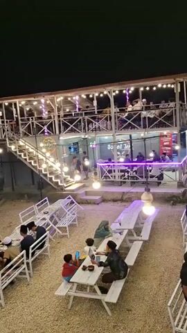 The video pans over an outdoor cafe at night. String lights illuminate the scene, highlighting several seating areas. In the foreground, a group of men in traditional Islamic attire sit at white picnic tables. To the left, several small, open-air bamboo huts are occupied by patrons. Further back, a multi-story building with an outdoor staircase is visible, also lit with string lights. People are seated at tables both on the ground level and on the upper floor of the building. The ground is covered with gravel.