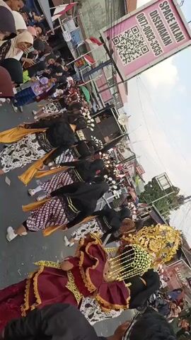 A group of people in traditional attire, possibly a dance troupe, are performing in a street. They are wearing dark tops and patterned skirts with sashes. Some individuals have ornate headpieces. A banner with a QR code and text in a foreign language is visible in the background, along with onlookers and parked motorcycles. The scene appears to be a cultural event or parade.