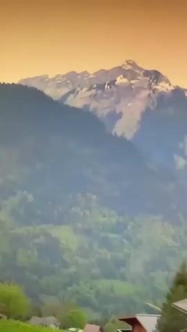 The video shows a panoramic view of a mountainous landscape with traditional wooden chalets nestled on green hillsides. The chalets are of various sizes, with dark wood exteriors and some featuring balconies. In the foreground, lush green fields with a white fence suggest a rural setting. In the background, a towering, rocky mountain peak rises sharply against a pale, warm-toned sky, with dense green coniferous forests covering the slopes. The overall impression is one of a serene and idyllic alpine village.