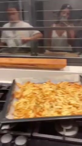 The video shows people preparing food in a kitchen. In the foreground, a sheet pan of french fries sits on the stovetop. A person is unwrapping raw steak on the counter.  Three people can be seen reflected in the window behind the counter.  One person is taking a selfie. There is a bottle of oil on the counter next to the french fries, and various items on the counter next to the steak.