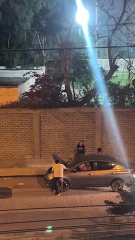 At night, a silver sedan is parked on a street with its hood open. A man in a white shirt is examining the engine while two people stand on the car near the windshield.  Two additional people are inside the car. A brick wall lines the edge of the street, and tall trees are visible behind it. A bright streetlight shines down on the scene.  Debris lies on the ground near the car.