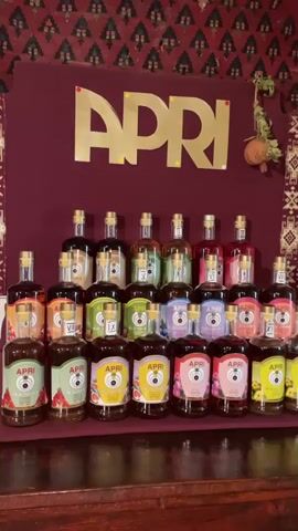 The video shows a display of APRi brand bottles, likely spirits or liqueurs, arranged in a tiered pyramid on a dark wooden surface. The bottles are of varying sizes and shapes, with distinct labels indicating different flavors. A large golden APRi logo is prominently displayed on a deep red background behind the bottles. The background also features a patterned fabric, possibly a tapestry or rug, with red and black geometric designs. A small, dry, bulbous object hangs on a vine to the right of the logo. The lighting is somewhat dim, casting a warm glow on the bottles. There are no people visible in the video.