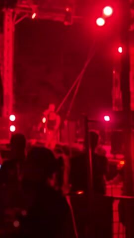The video shows a live music performance at night, indicated by the dark background and stage lighting. The stage is set up with large speakers, lighting rigs, and what appears to be musical equipment. A performer, dressed in dark clothing, is on stage, seemingly singing or rapping into a microphone. Several people are visible in the foreground, silhouetted against the bright red stage lights, suggesting they are part of the audience. The overall atmosphere is that of an energetic concert, with the red lighting creating a dramatic and intense mood. Some smoke or haze is also visible on stage, adding to the visual effect.