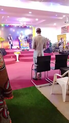 The video shows a church service in progress. Singers stand on a stage in front of a screen, backed by a band. A man stands in front of chairs in the aisle, facing the stage. A videographer stands on a platform, operating a camera. Another man stands next to the videographer. The floor is covered in red carpet, with a patch of artificial turf in front of the chairs.