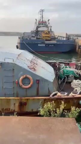 The video shows two boats docked at a harbor. The first boat is rusty and worn, with a life preserver visible on its side. It is a light blue-grey color with a sharply angled cabin. It has several round windows and the name "ALI 15" is partially visible on its side. The second boat, docked further away, is a larger blue tugboat named "MARIN K SEA". The water in the harbor appears calm, and the sky is overcast. There are harbor walls visible in the background.