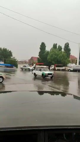 The video shows an outdoor scene on a cloudy day, possibly raining, with several vehicles present on a wet, reflective surface that appears to be a road or parking lot. In the foreground, the hood and windshield of a dark-colored vehicle are visible, reflecting the surroundings. In the middle ground, a white car with green accents is driving towards the right. Behind it, a silver car is also moving. Further back, there are several trucks, including a white truck with a crane. On the right side of the image, a modern, multi-story building with large glass windows is visible. The building has signage that reads "TAM İNSAAT". To the right of this building, there is a yellow construction vehicle. The overall environment appears to be an urban or semi-urban area.