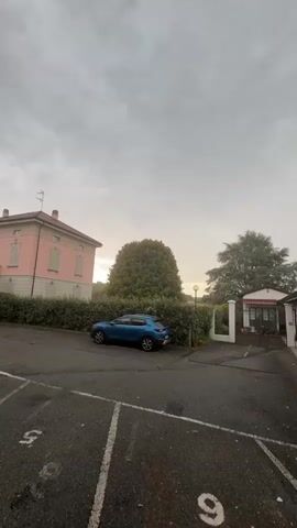 The media file shows a cloudy day in Sesto Calende, Italy. The scene is a parking lot in front of a building with a pink facade and white trim, possibly a residence or a small hotel. A blue SUV is parked in the lot. Behind the building is a dense hedge, and beyond that, a large, mature tree stands prominently. To the right of the large tree, there's a smaller building with a covered entrance and a light-colored fence. The sky is overcast, suggesting a gloomy or potentially rainy day. The time of day appears to be late afternoon or early evening, as indicated by the soft, diffused light. There are white painted lines on the asphalt marking parking spaces, with the numbers "5" and "9" visible on some of them. The overall mood is quiet and still.