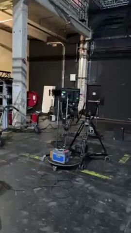 The video pans around a large, industrial space with a high ceiling and gray painted concrete floors. There are numerous pieces of film or video production equipment, including cameras, tripods, lighting stands, and a large camera crane. The crane is black with a long, adjustable arm. There are also some boxes and other miscellaneous items scattered around the space. At one point, a person walks by in the background wearing a dark jacket and face mask.
