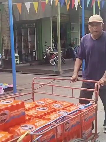 A man in a purple t-shirt and a tan baseball cap is pushing a red cart filled with orange-colored items. The cart appears to be a trolley with a metal frame. In the background, there are storefronts with shelves displaying items and a motorcycle parked in front of one of them. Colorful triangular flags are strung across the top of the scene, suggesting a festive or market atmosphere. The ground is paved. Another person, wearing a green t-shirt and dark shorts, walks past the storefronts in the background and appears to be holding a yellow object. The video cuts between these two scenes.