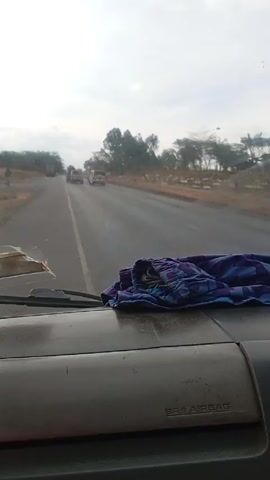 The video depicts a view from inside the cab of a vehicle, likely a truck, traveling on a paved road. The road has a center line and appears to be a major thoroughfare, with other vehicles, including trucks and cars, visible in the distance. Trees line the roadside, and the sky is overcast. On the dashboard in front of the camera is a piece of purple and blue patterned cloth. A person is walking along the side of the road, but it's unclear if they are on the shoulder or the road itself.