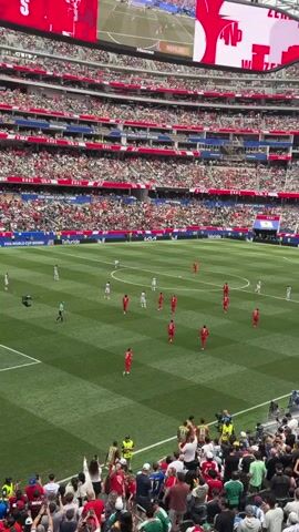 A wide-angle video captures a soccer match in a large, modern stadium. The stands are packed with spectators, most wearing red and white, suggesting they are supporting one of the teams. The field is a vibrant green with white markings. Players in red and white uniforms are visible on the field, engaged in play. A large video screen at the top of the stadium displays a replay of a past event. Above the field, the word "FIFA" is prominently displayed. The overall atmosphere appears to be energetic and festive, with fans actively watching the game.