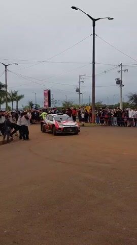 A white and red rally car with the number 34 visible on its side is seen in a dirt and asphalt area. The car is turning, with its tires kicking up dust. A crowd of people is gathered on the right side of the car, some holding up cameras. On the left side of the car, a few people are standing closer to the vehicle. The background shows palm trees, streetlights, utility poles with wires, and some buildings. The sky is overcast. The car appears to be participating in some sort of event, possibly a rally or a demonstration, with spectators present.