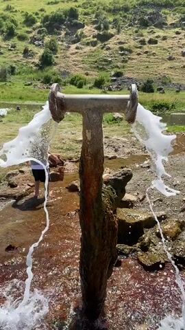 A boy walks barefoot in shallow water where two streams of water pour out of a rusty metal pipe that is vertically installed in the ground. The pipe is shaped like a T with two flanges at the top. The boy is wearing shorts and a t-shirt. The area around the pipe is rocky and the water is reddish-brown. In the background, there is a grassy hill with some trees and a body of water that appears to be a river. The sky is not visible in the frame.
