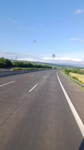 The video shows a view from a vehicle traveling on a highway. The highway is paved with asphalt and has multiple lanes separated by white lines. The road curves to the right in the distance. On the left side of the road, there is a concrete barrier and then a line of green trees. On the right side of the road, there is a metal guardrail and then a field of yellow crops. The sky is blue with a few scattered white clouds. There are some dark circular spots visible in the footage, possibly from the camera lens. A few distant vehicles can be seen on the highway.