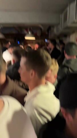 The video depicts a crowded, dimly lit bar interior, filled with people standing and sitting. In the foreground, the back of multiple heads are visible, including a blonde woman with a black blazer and a man with a grey hoody. The focus shifts to a group seated at a wooden table with beer glasses and shot glasses. One man wearing a green hoodie and a baseball cap has his arm around one of the woman at the table, and appears to be taking a picture of them with his phone. A vintage radio, a cash register, and various stickers can be seen behind the seated group. The video captures the lively atmosphere, showing people engaging in conversation, moving around, and socializing. The environment is compact, with low ceilings and visible fixtures and elements like the ventilation system.