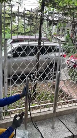 A person wearing a blue long-sleeved shirt, yellow pants, and black gloves is welding a metal fence. The person is holding a welding torch and is welding a vertical metal post. There is a bright spark of light from the welding. The fence is made of metal with a diamond pattern. Behind the fence, there is a silver car parked. Trees and foliage are visible in the background. The scene appears to be outdoors.