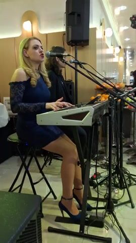 Two women are playing a keyboard and singing at a venue, likely a restaurant. The woman on the left is wearing a blue dress and high heels and singing into a microphone. The woman on the right is playing the keyboard, also with a microphone. A speaker, microphone stands, cables, and other equipment are nearby.  A buffet-style table of oranges can be seen behind the musicians. The lighting changes slightly from green to blue.