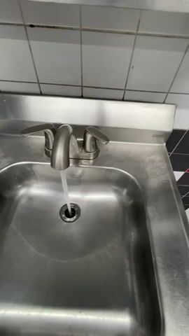The video shows a close-up view of a stainless steel sink with water running from the faucet into the drain. The faucet is a brushed nickel finish with two handles. The sink is clean, but there are some water spots visible. The background shows a tiled wall and a black and white tiled backsplash. The water stream is steady and appears to be cold. The video is shot from a slightly elevated angle, looking down into the sink.