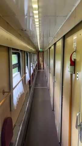 The video shows the interior of a train car hallway. The hallway is narrow, with a row of windows on the left and cabin doors on the right. The walls are wooden paneling, and the floor is a dark brown. The ceiling has a row of bright fluorescent lights running the length of the hallway. Small, white curtains hang in the windows. There's a red emergency handle mounted on the wall near the beginning of the hallway. The train is in motion.
