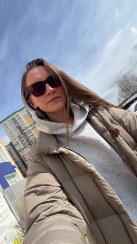 The video shows a woman taking a selfie with a baby in a stroller. The woman has long brown hair and is wearing sunglasses, a gray hoodie, and a beige puffer jacket. She smiles at the camera and then laughs while pushing the stroller. The baby is wearing a yellow beanie and green sunglasses, and is lying in the stroller. The background of the video shows a street with parked cars and buildings. The sky is blue with some clouds.