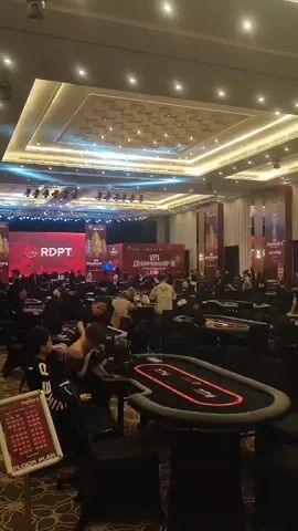 The video shows a large room filled with people playing poker at multiple tables. The room is well-lit with a patterned carpet and a decorative ceiling. There's a stage with large screens displaying the tournament name "RDPT" and "VPT CHAMPIONSHIP III". The poker tables are black with red trim. People are seated around the tables, some watching, others actively playing. A floor plan of the event is visible on a stand near the camera. The atmosphere appears to be focused and competitive.