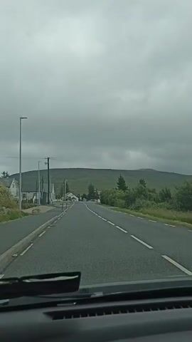 This is a first-person perspective video taken from inside a vehicle, likely a car, as it drives along a paved road. The road is two-lane with a dashed white line marking the center. On the left side of the road, there is a sidewalk, a lamppost, a utility pole with wires, and several buildings that appear to be houses or small businesses. There are also some trees and bushes lining the road. In the distance, a large, green, rolling hill or mountain dominates the landscape. The sky is overcast with heavy gray clouds, suggesting a cloudy or potentially rainy day. The interior of the vehicle is visible at the bottom of the frame, including the windshield wipers and part of the dashboard. The video appears to be a short, continuous recording with no discernible actions or interactions occurring other than the movement of the vehicle.