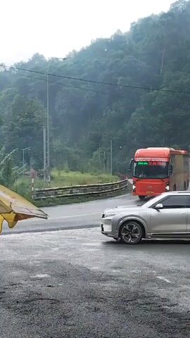 An orange and yellow bus with Vietnamese text is parked on the right side of a wet asphalt road. A silver SUV is parked in front of the bus, facing to the right. In the background, a white van is driving on the road, away from the camera. The road is surrounded by lush green trees and a misty mountain in the distance. The sky is overcast and gray. A yellow tarp is visible on the left side of the frame.