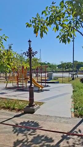 The video pans across a park area on a sunny day. A lamppost is in the foreground, casting a shadow. Beyond it, a playground with swings, a slide, and climbing structures is visible. Trees line a paved walkway. In the background, there are more trees, a blue sky, and some structures that could be buildings or utility towers.