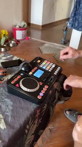 A close-up, medium shot focuses on a person's hands operating a black DJ controller. The controller has a circular jog wheel, numerous colorful buttons, and a small digital display screen. The person's fingers are actively pressing buttons on the right side of the controller. The controller is placed on a table covered with a patterned tablecloth. To the left of the controller, a smartphone is visible, as well as some colorful, reflective objects, possibly party lights. In the background, a doorway or hallway is visible, with wooden flooring and some items on the floor including a white bucket, some tools, and a partially visible person wearing a grey slipper. The lighting appears to be natural, coming from an unseen source.