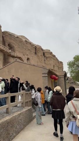 The video shows a group of people standing on a path behind a low fence, looking at a large tan rock formation that looks like a cliff. The rock face has several small buildings built into it, some with wooden walkways connected to the cliff face. Some people are taking photos of the scene. The weather is overcast. There are a few red lanterns hanging on a building. There are stone stairs leading up towards the buildings in the rock face.