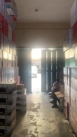 The video shows a narrow hallway or aisle in a warehouse, with stacks of boxes lining both walls. The boxes appear to be filled with electronics, possibly printers, based on the visible packaging. Sunlight streams in from an open doorway at the end of the hallway, creating a bright silhouette of a person sitting on a bench. Another person is partially visible outside the doorway, near a motorcycle. The floor is concrete and appears wet or stained in places. The overall impression is one of a storage area within a commercial or industrial setting.