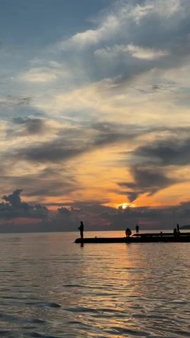 The video shows a sunset over a calm body of water. Silhouettes of people are visible on a pier or dock in the foreground, some of whom appear to be fishing. The sky is filled with dramatic clouds, colored orange, yellow, pink, and blue by the setting sun. The water reflects the colors of the sky.